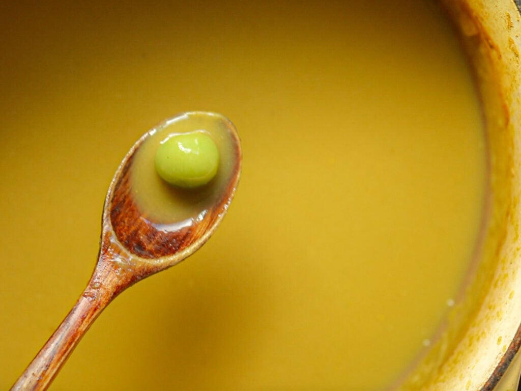 A wooden spoon hovers over a bowl of smooth, creamy broccoli soup, holding a single green rice dumpling. The soups surface is reflective and looks rich and thick. The scene is close-up, focusing on the contrasting textures and colors.